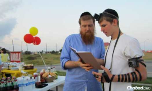 The rabbi helps Jacob Feldman don tefillin at an outdoor barbecue for the community.