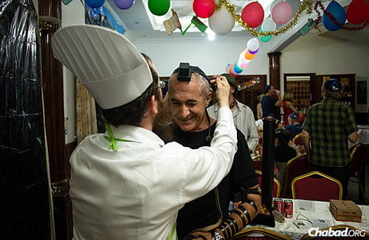 A guest puts on tefillin in Cambodia last year at “Purim in Italy” and joins in a festive meal, the final mitzvah associated with the holiday.