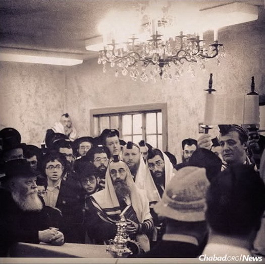 The Rebbe, left, and worshippers at 770 Eastern Parkway look on as Jerry Weintruaub, right, is given the aliyah of hagbah, raising the Torah scroll at the completion of the mid-week reading.